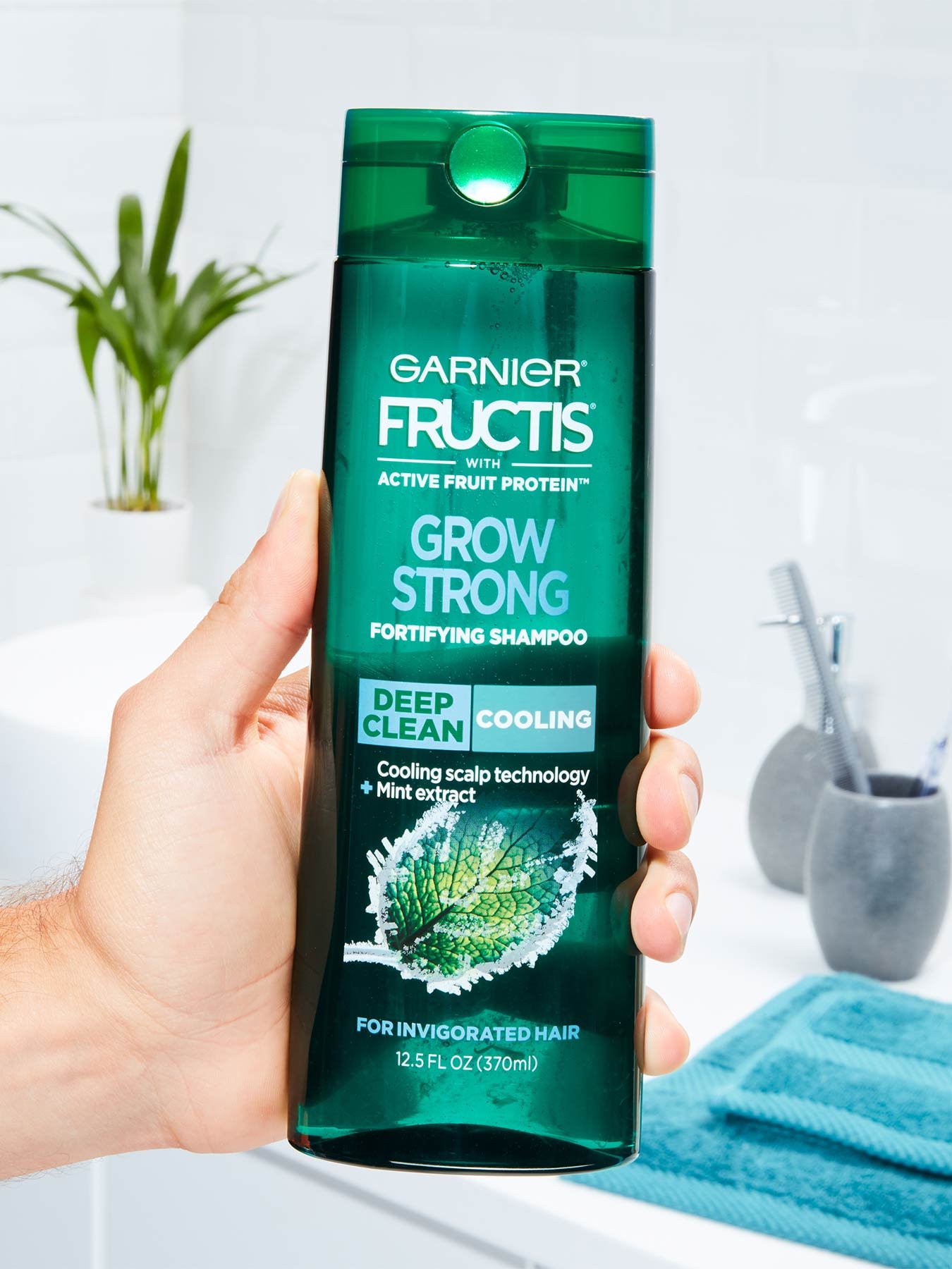 Hand holding Cooling Deep Clean Shampoo for Men for Invigorated Hair in a white bathroom with a blue towel, silver cups with toothbrushes, and a plant.